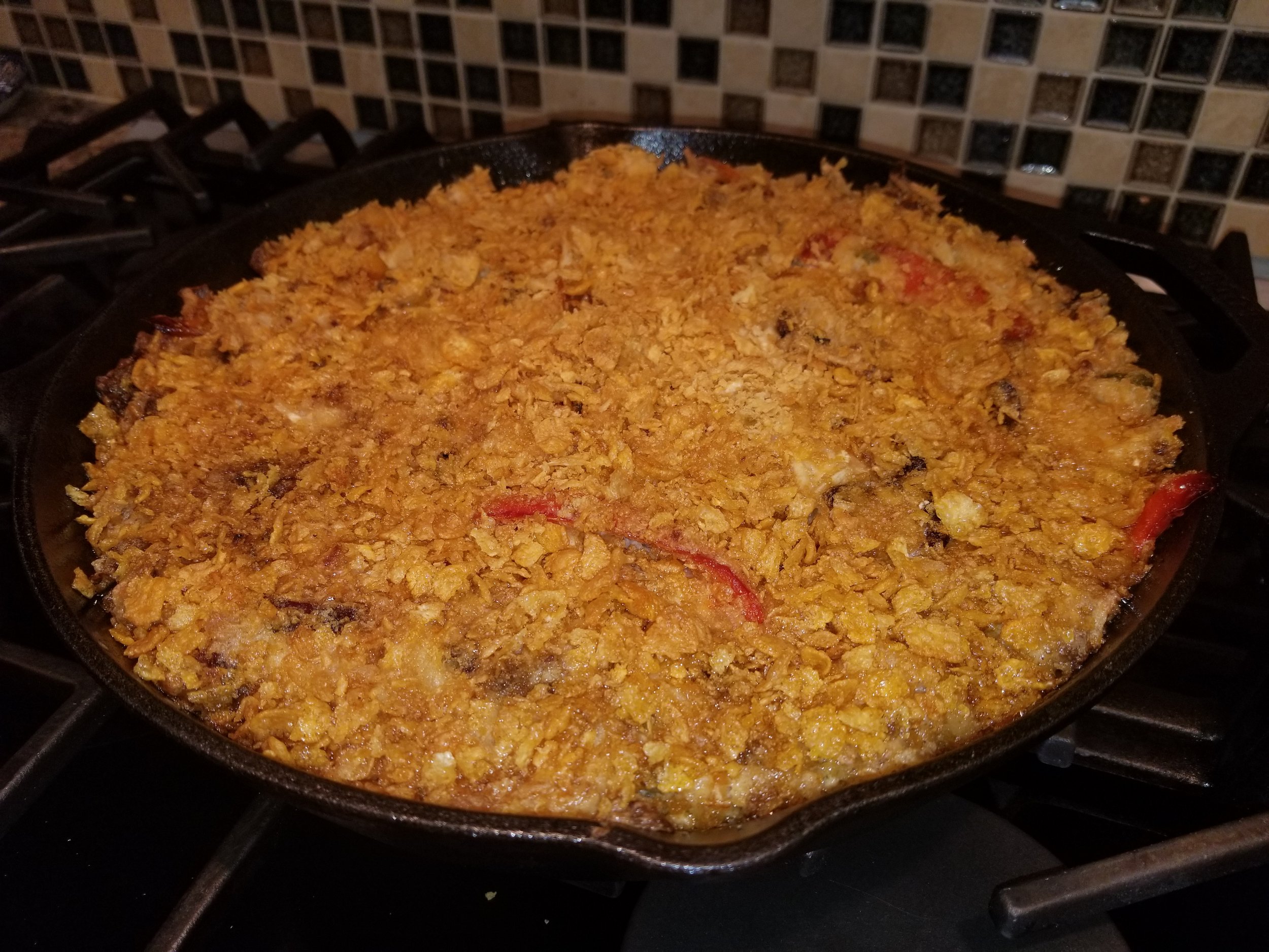 Crispy baked casserole in a cast iron skillet, topped with golden corn flakes and featuring colorful bell peppers, resting on a gas stove.
