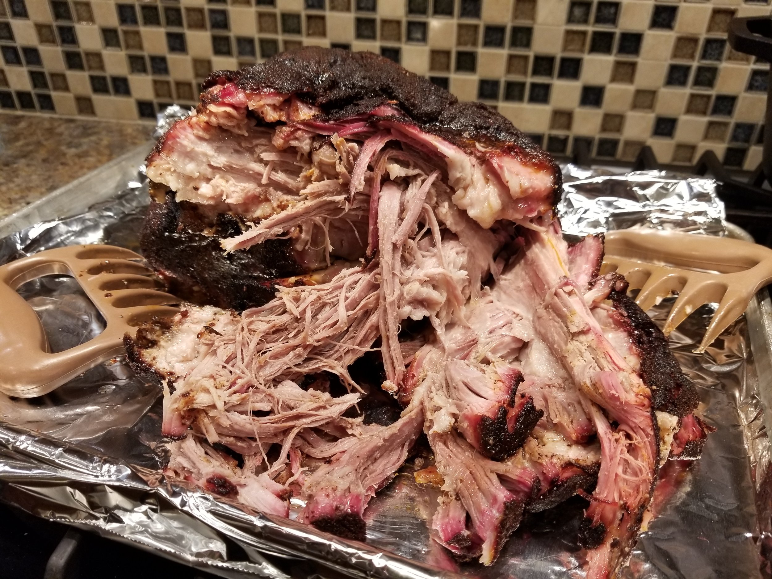 Pulled pork with a smoky, charred exterior, showcasing tender, shredded meat on a foil-lined tray, accompanied by meat claws for serving. Ideal for BBQ enthusiasts and recipes.