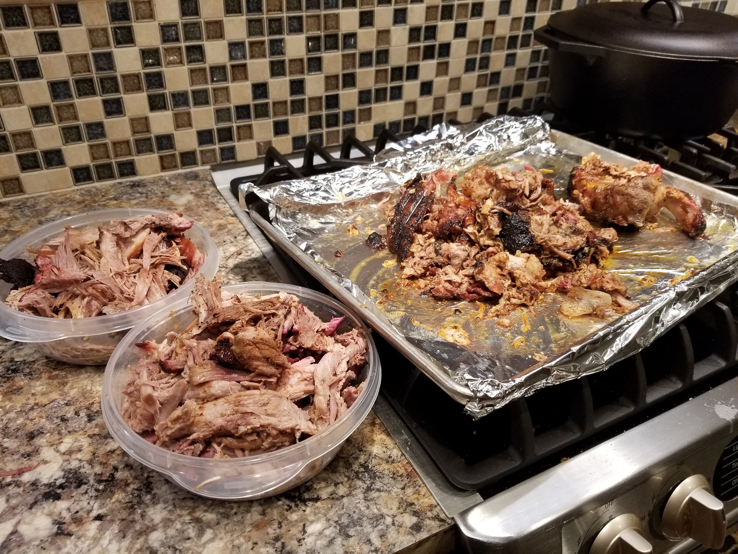 Pulled pork served in two clear containers alongside a foil-lined baking sheet with leftover meat, set on a granite countertop near a stovetop.