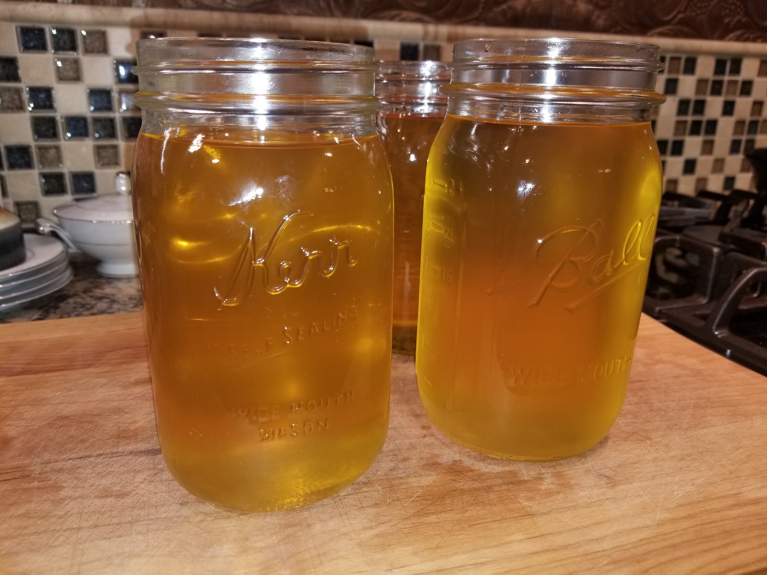 Three glass jars filled with golden liquid are displayed on a wooden countertop, with a textured backsplash in the background. The jars are labeled Kerr and Ball, indicating they are mason jars, commonly used for canning and preserving.