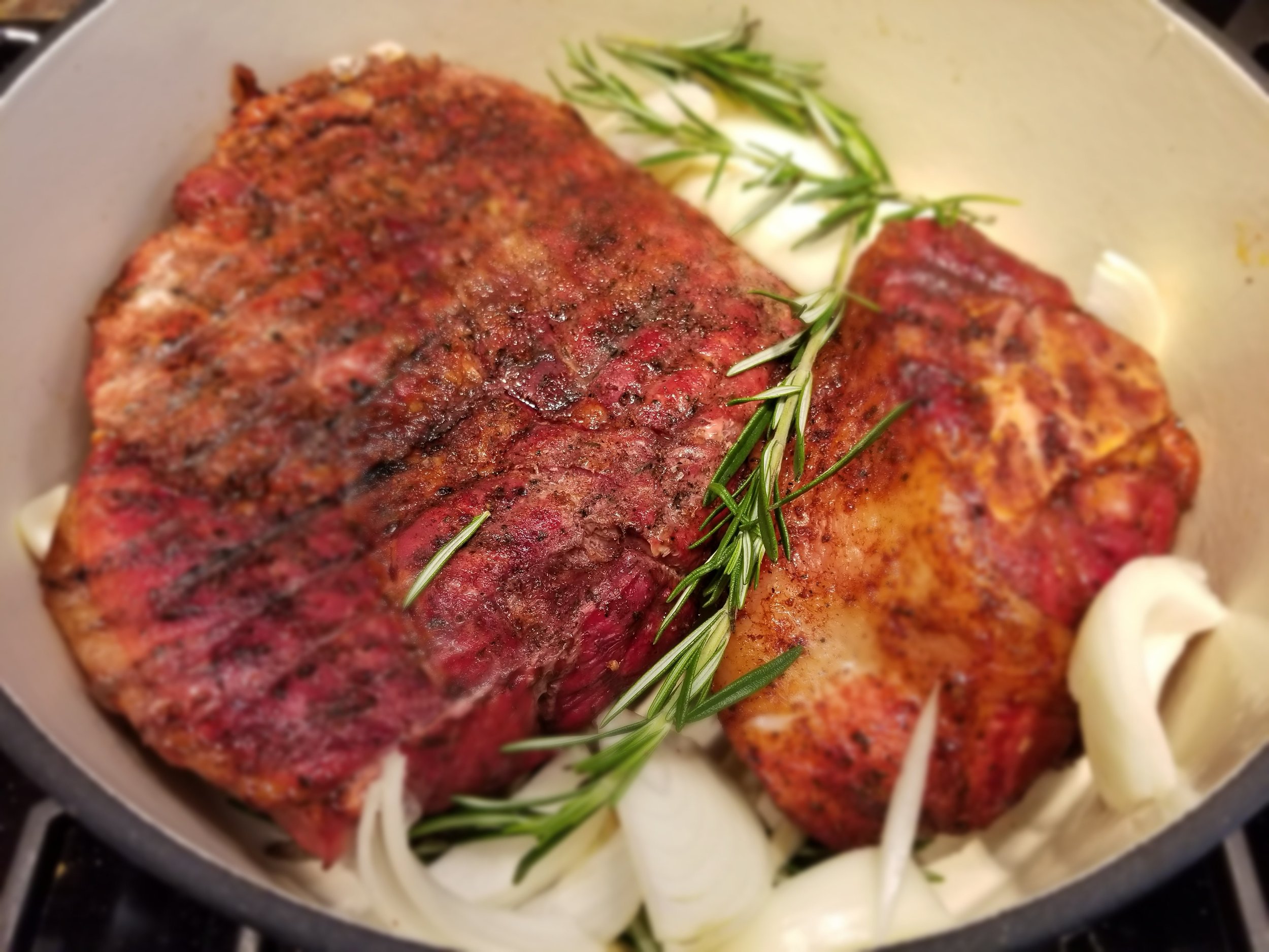 Grilled beef cuts resting on a bed of sliced onions and fresh rosemary in a cooking pot, showcasing a flavorful preparation for a hearty meal.