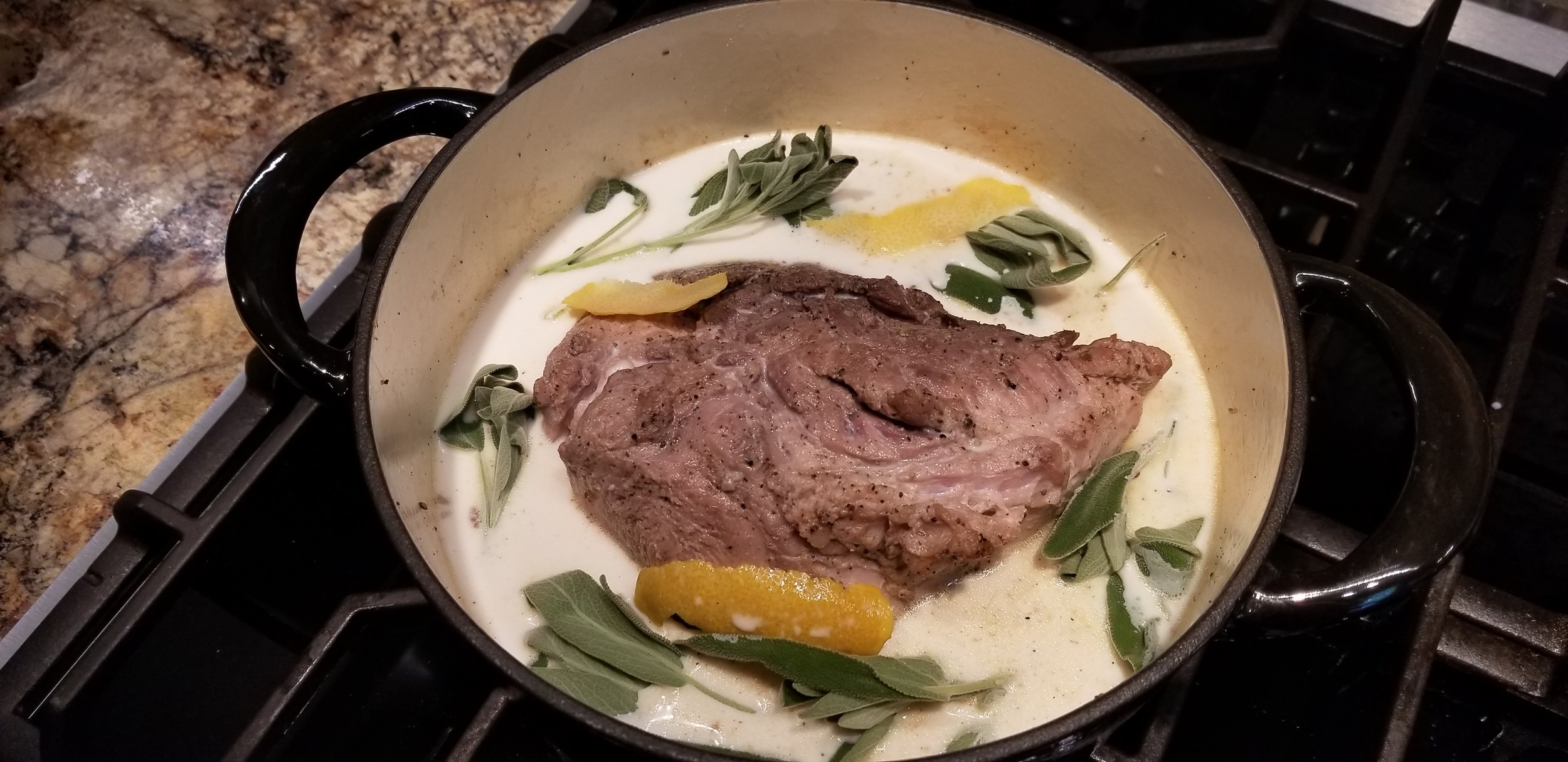A pot on a stove containing a large piece of meat simmering in a creamy sauce with fresh sage leaves and lemon zest, showcasing a delicious cooking preparation.