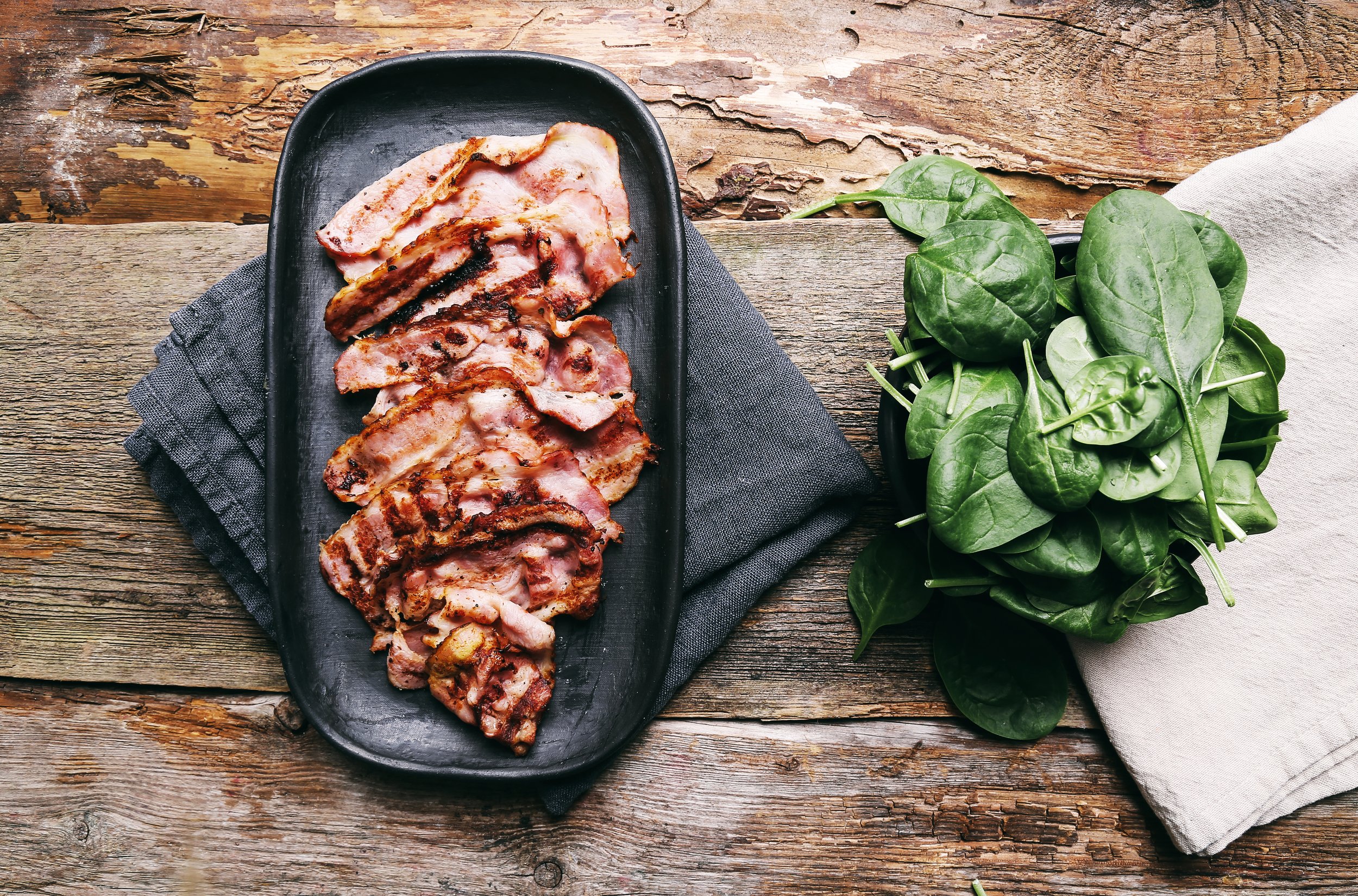 Sliced crispy bacon on a black platter beside fresh spinach leaves on a wooden table, showcasing a delicious breakfast option.