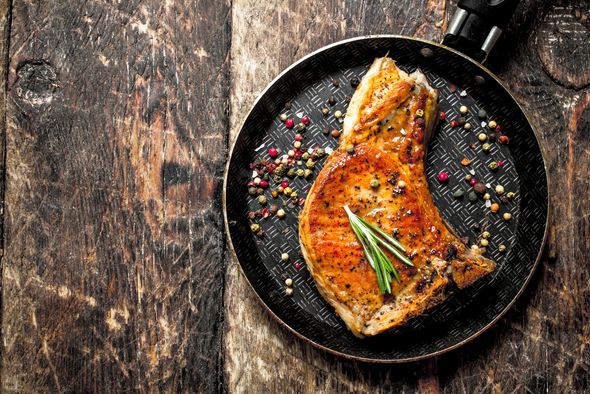Grilled pork chop seasoned with black pepper and garnished with fresh rosemary, served on a textured black plate with colorful peppercorns, set against a rustic wooden background.