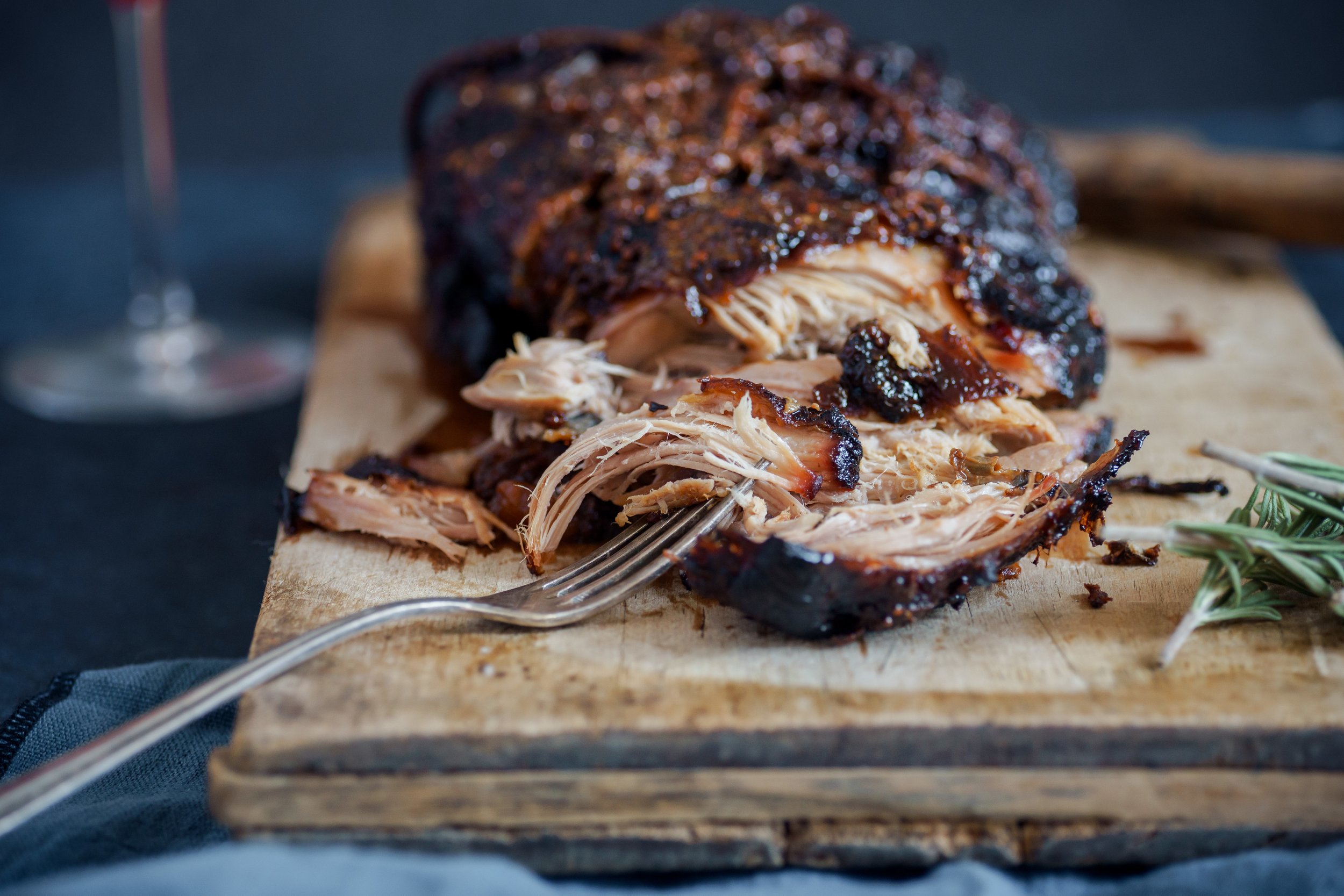 Close-up of tender, pulled smoked pork on a wooden cutting board, garnished with fresh rosemary, showcasing its juicy texture and rich, caramelized exterior. A fork is placed beside the meat, hinting at a delicious meal.