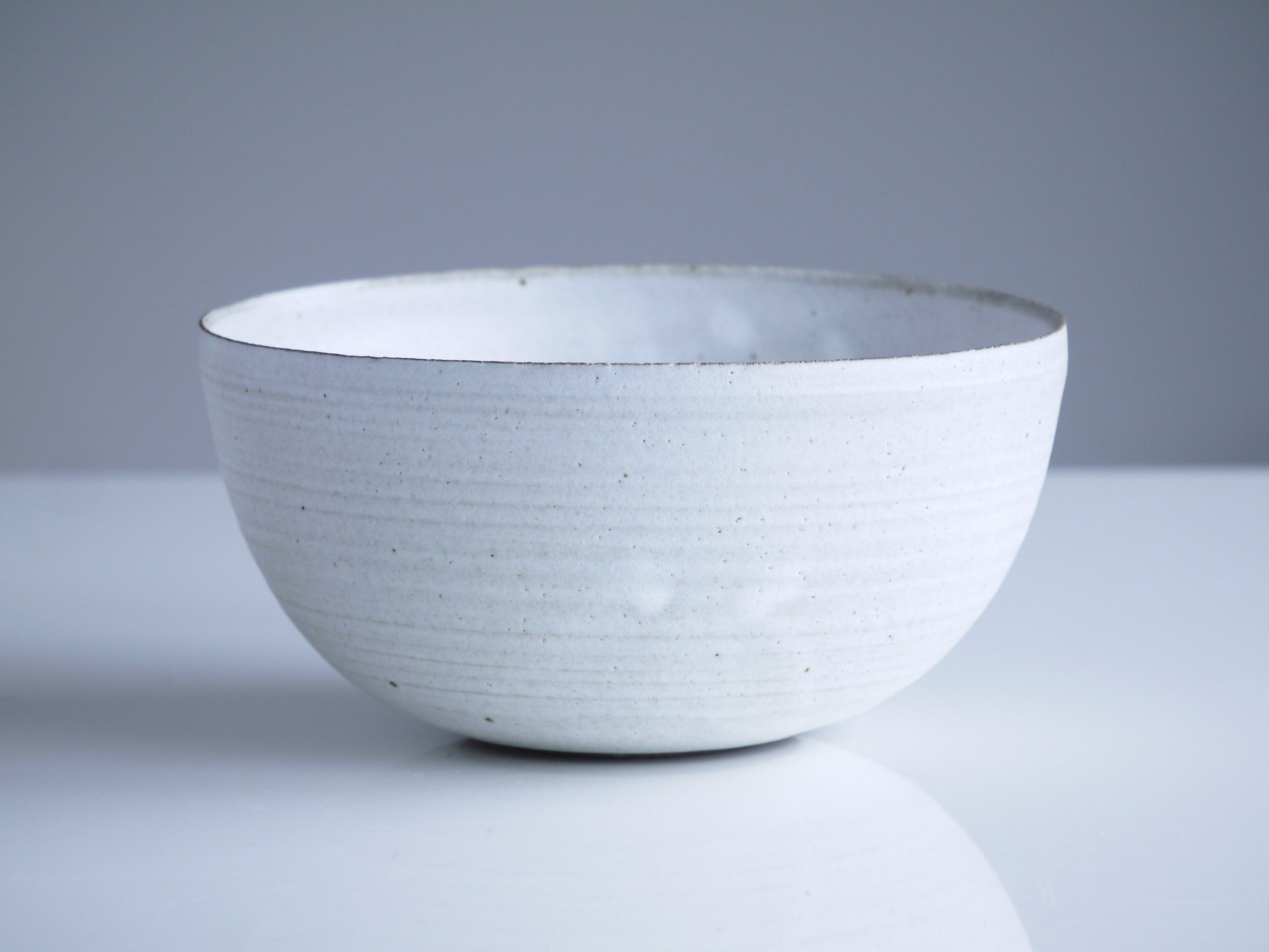 Ceramic bowl with a simple white glaze and subtle textured lines, set against a soft gray background, ideal for serving or decorative use.