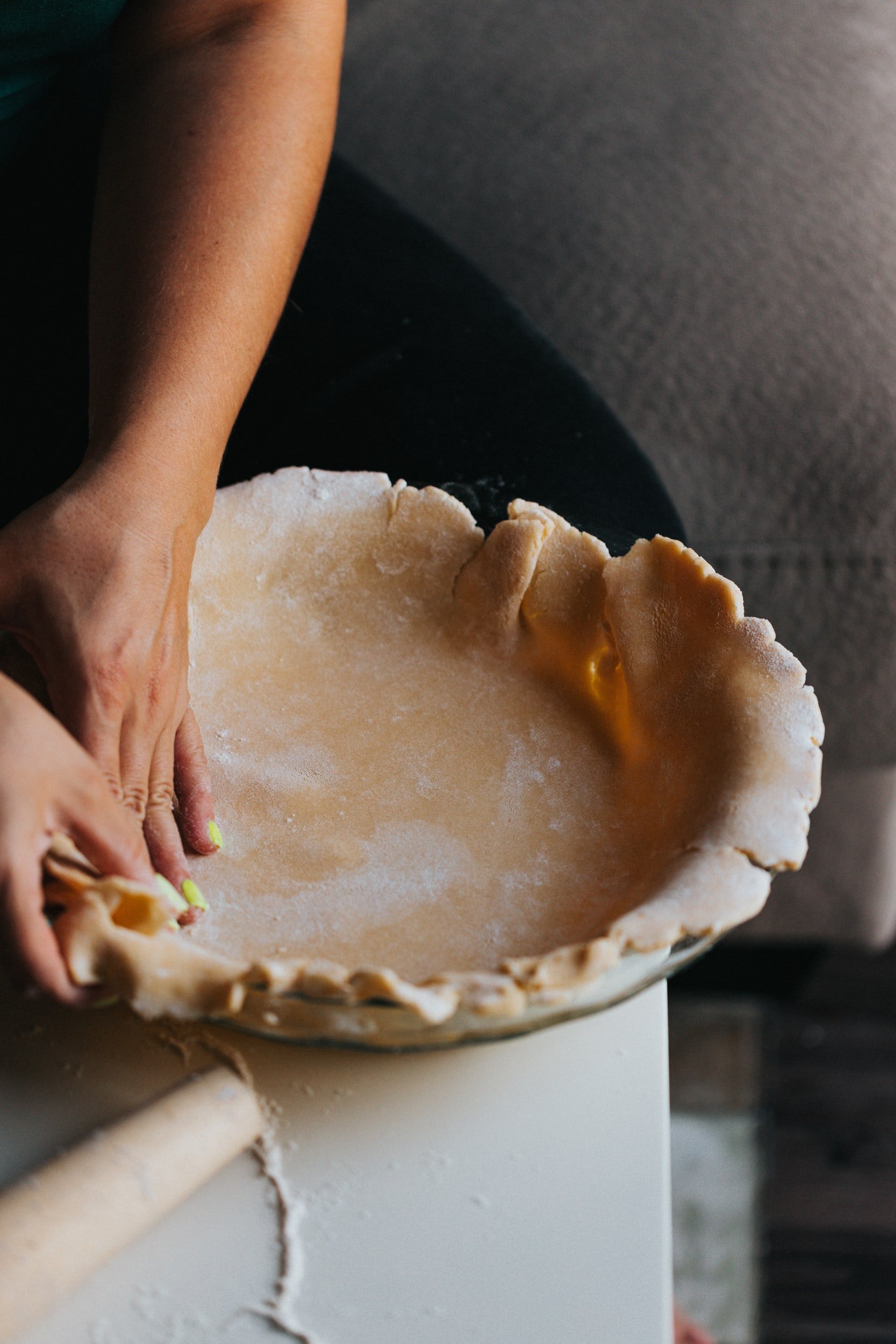 Person preparing a homemade pie crust, gently shaping the edges of the dough in a glass pie dish, with a rolling pin and flour scattered on the table.